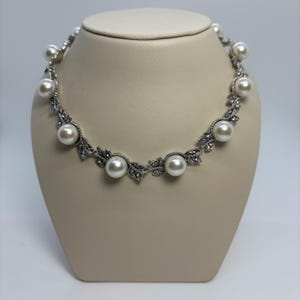 May include: A silver necklace with large, round, white pearls and leaf-shaped details. The necklace is displayed on a cream mannequin, highlighting its design. This piece of jewellery is designed to be worn around the neck.