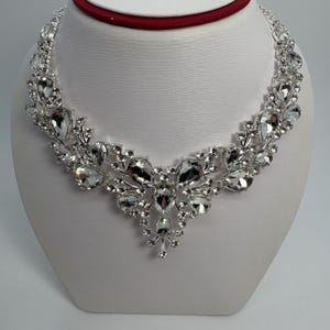 May include: A silver-toned necklace with clear, sparkling crystals. The necklace features a symmetrical design with large, teardrop-shaped crystals and smaller, round stones. It is displayed on a white bust, showcasing its elegant and glamorous style.