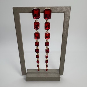 Puede incluir: Pendientes largos y colgantes con una serie de piedras preciosas rojas rectangulares graduadas. Las piedras están conectadas por eslabones de metal dorado. Los pendientes se exhiben en un soporte gris sobre un fondo blanco.