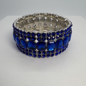 May include: A silver-toned stretch bracelet featuring rows of vibrant blue rectangular and round gemstones. The bracelet has a detailed, openwork design on the metal band, creating a sparkling and elegant accessory.