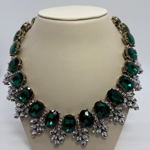 Peut inclure: Un collier ras de cou avec de grandes pierres ovales de couleur vert émeraude et de petits strass transparents. Le collier est de couleur or.