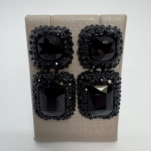 Może przedstawiać: Para czarnych, kwadratowych kolczyków wiszących. Każdy kolczyk ma dwa kwadratowe, fasetowane, czarne kamienie. Kamienie są otoczone mniejszymi, czarnymi, błyszczącymi kamieniami. Kolczyki są prezentowane na neutralnym, pokrytym tkaniną stojaku.