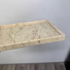 Op de afbeelding: Een rechthoekige marmeren tray met een opstaande rand. De tray heeft een beige en bruin gemarmerd patroon. De tray wordt waarschijnlijk gebruikt voor decoratieve doeleinden of om kleine items in te bewaren.
