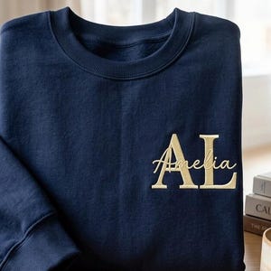 May include: A navy blue crewneck sweatshirt featuring embroidered text on the front left chest. The design includes the large capital letters AL in a serif font, with the name Amelia written in a cursive script layered over the letters.