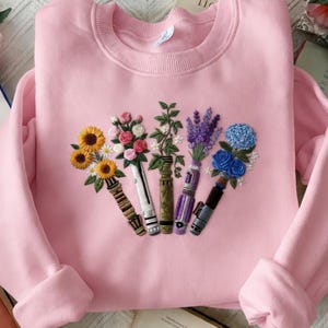 Puede incluir: Una sudadera rosa claro con dise&ntilde;os florales bordados. El bordado presenta girasoles, rosas, lavanda y flores azules dispuestas en un patr&oacute;n &uacute;nico. La sudadera tiene mangas largas y cuello redondo.