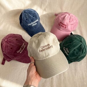 May include: Five baseball caps in various colors: navy blue, pink, burgundy, green, and tan. Each cap features embroidered text: "Daydreaming Often," "Always Up For It," "Good Times Advocate," "Stay Curious," and "Custom Embroidery."