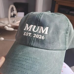May include: A green baseball cap with the word "MUM" embroidered in white, along with "EST. 2026". The cap has a curved brim and a classic six-panel design.