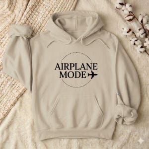 May include: A beige hoodie with a front pocket and a hood. The front of the hoodie features the words "AIRPLANE MODE" in a black, circular design, with a small airplane graphic. The hoodie is made of a soft material.