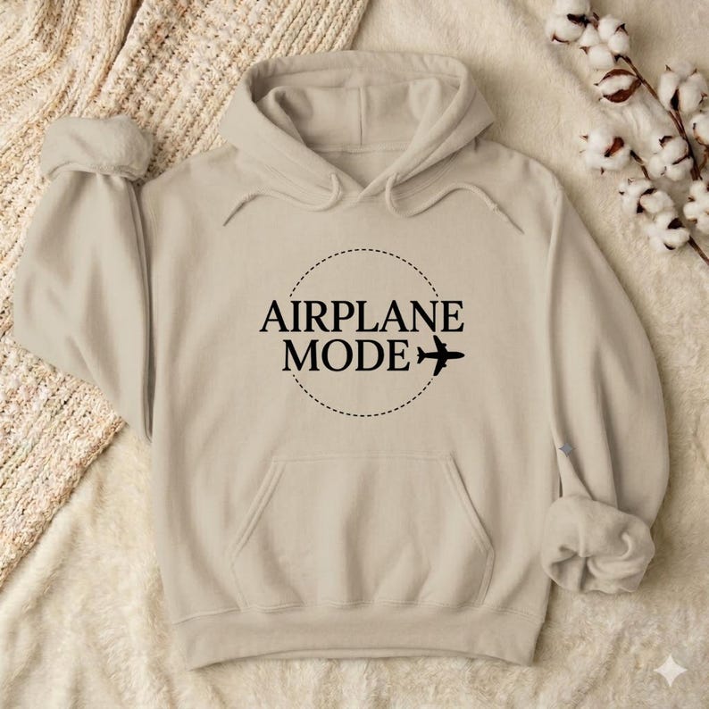 May include: A beige hoodie with a front pocket and a hood. The front of the hoodie features the words "AIRPLANE MODE" in a black, circular design, with a small airplane graphic. The hoodie is made of a soft material.