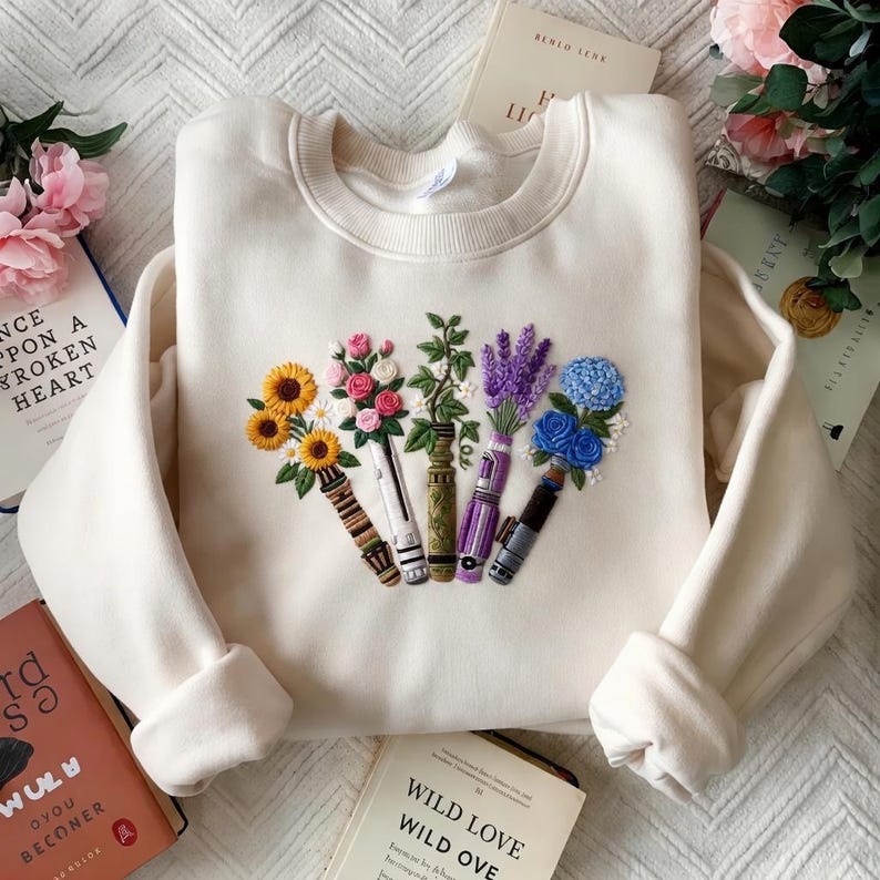 Puede incluir: Sudadera color crema con un dise&ntilde;o floral bordado. El dise&ntilde;o presenta girasoles, rosas, lavanda e hortensias azules dispuestas en fila. La sudadera tiene cuello redondo y mangas largas.