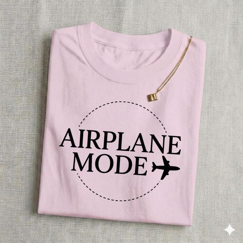 May include: A light pink t-shirt with the words "AIRPLANE MODE" in black, encircled by a dotted line, and a small airplane graphic. A gold necklace with a small pendant is draped over the shirt.