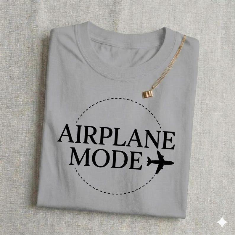 May include: A light gray t-shirt with the words "AIRPLANE MODE" in black, encircled by a dashed line, and a small airplane graphic. A gold necklace is draped over the shirt.