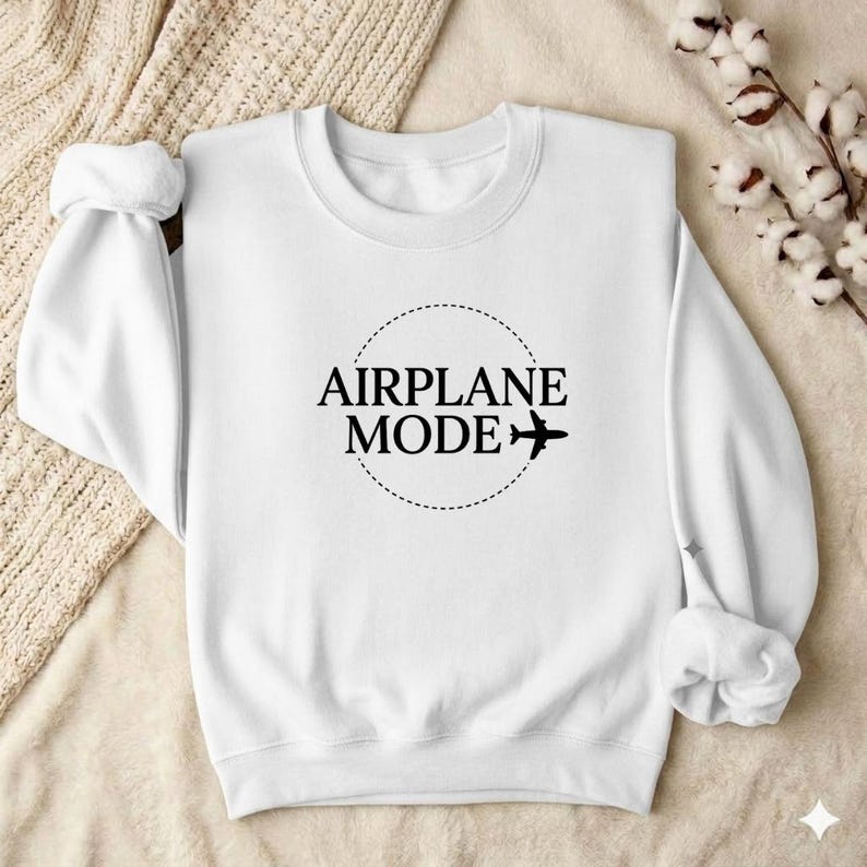 May include: White sweatshirt with the text "AIRPLANE MODE" in a black, circular design with a dotted border and a small airplane graphic. The sweatshirt has long sleeves with rolled-up cuffs.