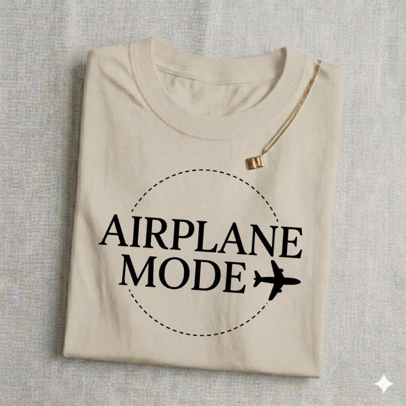 May include: Beige t-shirt with the words "AIRPLANE MODE" in black, inside a dotted circle, and a small airplane graphic. A gold necklace with a pendant is draped over the shirt.