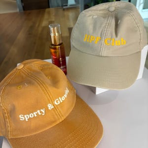 May include: Two baseball caps, one mustard yellow and one beige. The mustard cap has the text "Sporty & Glowing" in white. The beige cap has the text "HPF Club" in yellow. A bottle of Dove hair oil is also in the image.