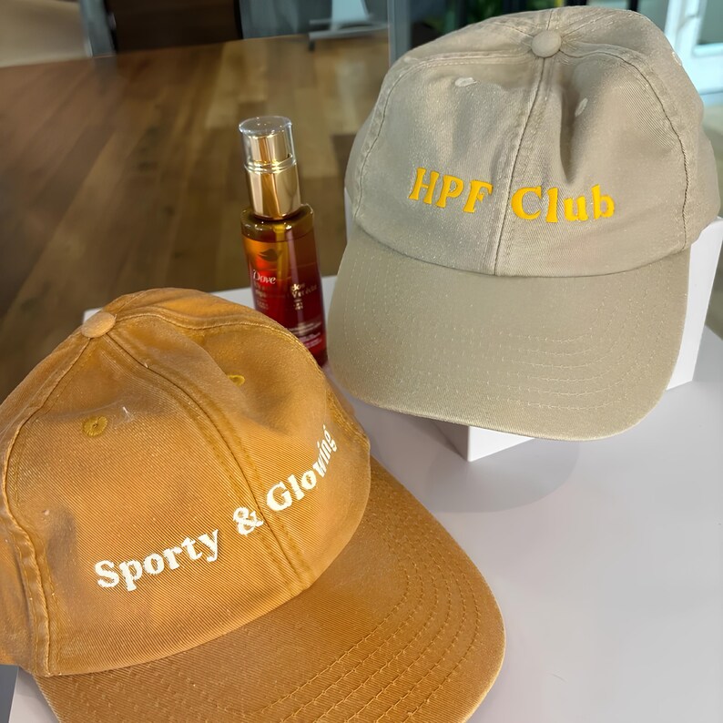 May include: Two baseball caps, one mustard yellow and one beige. The mustard cap has the text "Sporty & Glowing" in white. The beige cap has the text "HPF Club" in yellow. A bottle of Dove hair oil is also in the image.