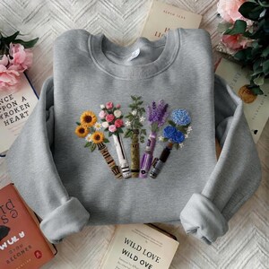 Puede incluir: Una sudadera gris con dise&ntilde;os florales bordados. El bordado presenta girasoles, rosas, lavanda y flores azules dispuestas en un dise&ntilde;o en forma de jarr&oacute;n. La sudadera tiene mangas largas y cuello redondo.