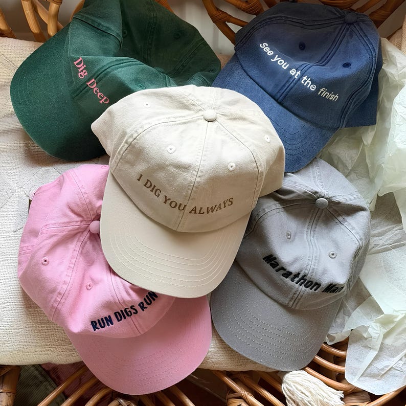 May include: A collection of baseball caps in various colors, including green, blue, pink, beige, and gray. Each cap features embroidered text such as "Dig Deep," "See you at the finish," and "Marathon Man."
