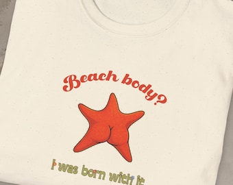 May include: Cream-colored t-shirt with the text "Beach body? I was born with it" above a red starfish graphic. The starfish graphic has a humorous design. The shirt is folded on a gray surface.