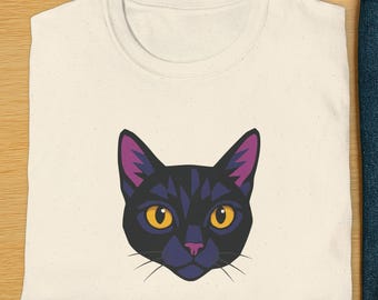 May include: Cream-colored t-shirt featuring a stylized black cat face with yellow eyes and purple accents. The cat graphic is centered on the shirt. Sunglasses, blue jeans, and white sneakers are also in the image.