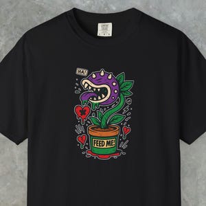 Puede incluir: Camiseta negra con un diseño de planta de dibujos animados. La planta tiene una cabeza morada, dientes puntiagudos y una lengua larga. Está en una maceta marrón con "FEED ME!" escrito, rodeada de corazones. Un bocadillo dice "HA!"