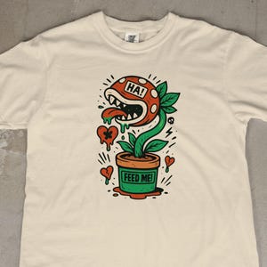 Puede incluir: Camiseta color crema con una ilustración de dibujos animados de una planta carnívora en una maceta. La planta tiene una cabeza roja con la palabra "HA!" y una lengua larga. La maceta dice "FEED ME!" con corazones y rayos.