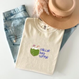 May include: Cream-colored t-shirt with a green coconut graphic and the words "Chillin' and Sippin'" in blue. The shirt is paired with light blue jeans, a tan hat, and a necklace. The image has a casual, summery aesthetic.