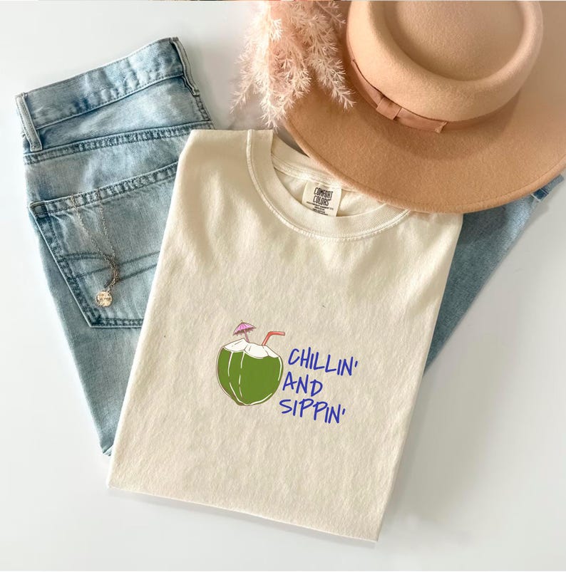 May include: Cream-colored t-shirt with a green coconut graphic and the words "Chillin' and Sippin'" in blue. The shirt is paired with light blue jeans, a tan hat, and a necklace. The image has a casual, summery aesthetic.