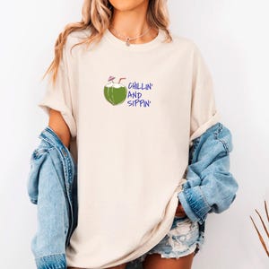 May include: Cream-colored t-shirt with a green coconut illustration and the words "CHILLIN' AND SIPPIN'" in blue. The shirt is paired with a denim jacket and shorts, creating a casual, summery look.