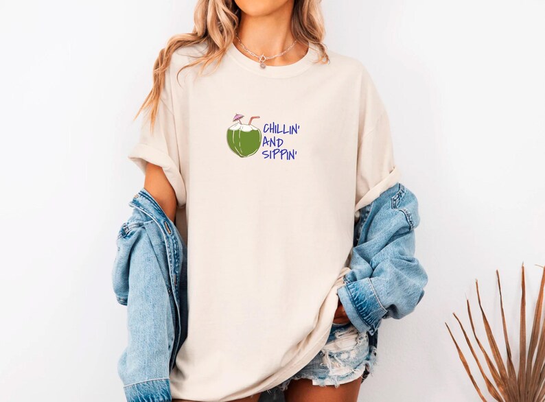May include: Cream-colored t-shirt with a green coconut illustration and the words "CHILLIN' AND SIPPIN'" in blue. The shirt is paired with a denim jacket and shorts, creating a casual, summery look.