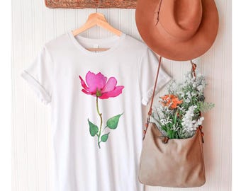 May include: White t-shirt with a pink flower graphic. The flower has green leaves and a long stem. The shirt is hanging on a wooden hanger.