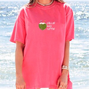 May include: A coral-colored t-shirt with a graphic of a coconut with two straws and the words "Chillin' and Sippin'" in white. The shirt has short sleeves and a relaxed fit, perfect for a day at the beach.
