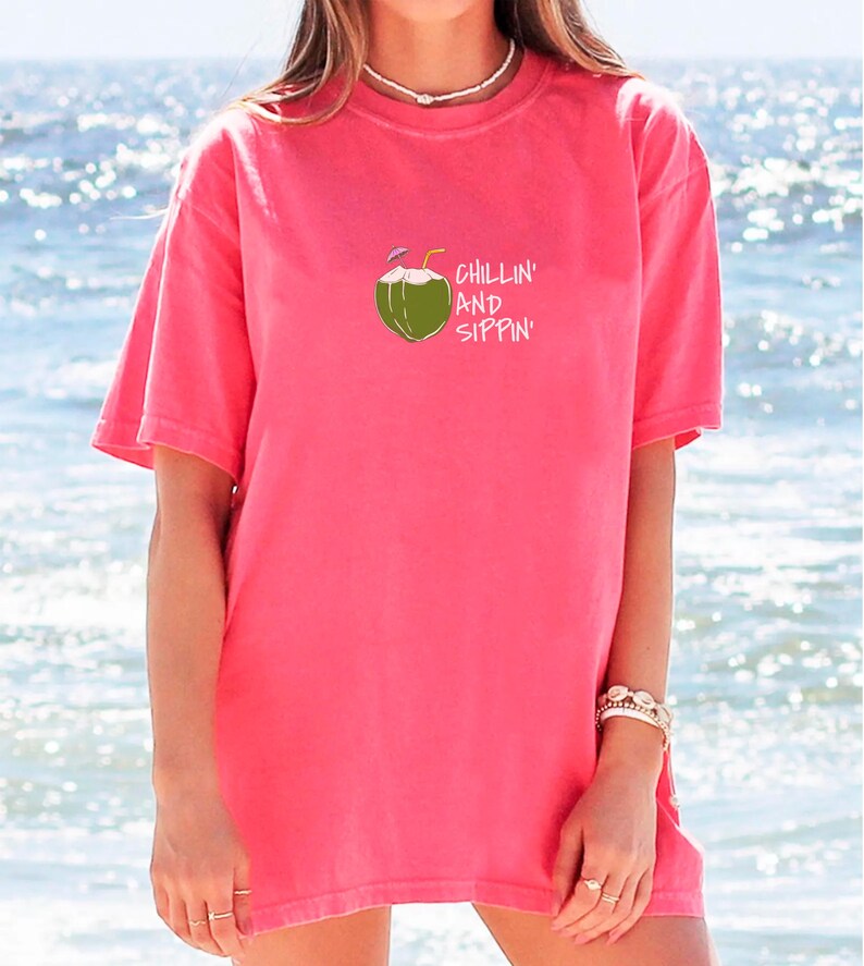 May include: A coral-colored t-shirt with a graphic of a coconut with two straws and the words "Chillin' and Sippin'" in white. The shirt has short sleeves and a relaxed fit, perfect for a day at the beach.