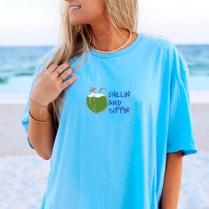 May include: Light blue t-shirt with a graphic of a green coconut with two straws and the words "Chillin' and Sippin'" in blue. The shirt is made of a soft material and is perfect for a day at the beach.
