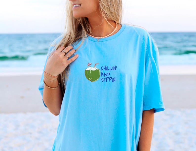 May include: Light blue t-shirt with a graphic of a green coconut with two straws and the words "Chillin' and Sippin'" in blue. The shirt is made of a soft material and is perfect for a day at the beach.