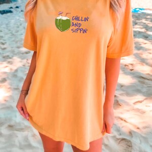 May include: Mustard yellow t-shirt with a graphic of a green coconut with a straw and the words "CHILLIN' AND SIPPIN'" in blue. The shirt is short-sleeved and appears to be made of a soft material. The model is standing on a sandy beach.
