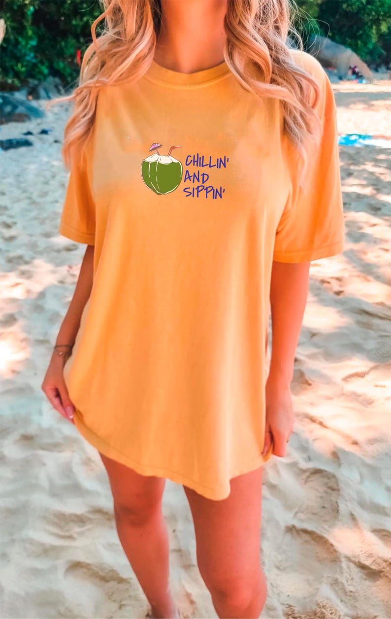 May include: Mustard yellow t-shirt with a graphic of a green coconut with a straw and the words "CHILLIN' AND SIPPIN'" in blue. The shirt is short-sleeved and appears to be made of a soft material. The model is standing on a sandy beach.