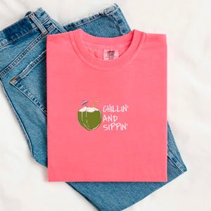 May include: A coral-colored t-shirt with a crew neck and the words "CHILLIN' AND SIPPIN'" printed on the front. The graphic features a green coconut with a straw and a small umbrella. The shirt is laid on top of blue denim jeans.