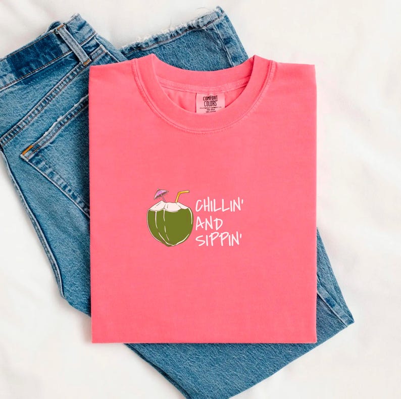 May include: A coral-colored t-shirt with a crew neck and the words "CHILLIN' AND SIPPIN'" printed on the front. The graphic features a green coconut with a straw and a small umbrella. The shirt is laid on top of blue denim jeans.
