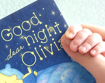 May include: A children's book titled "Good Night Dear Olivia" with a dark blue cover and gold lettering. The book features an illustration of a child reading. A small hand holds the book.