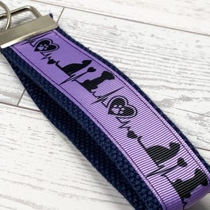 May include: A navy blue wristlet keychain with a purple ribbon featuring black silhouettes of dogs, paw prints, and a heartbeat design. The keychain has a silver metal clasp.