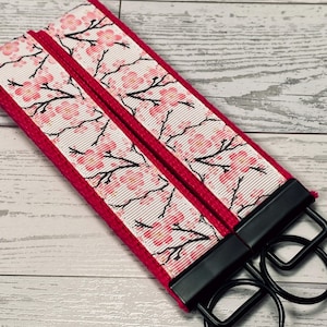 May include: Two key fobs with a cherry blossom design. The key fobs have a white ribbon with pink cherry blossoms and black branches. The edges are a solid red. The key fobs have black metal hardware and black key rings.