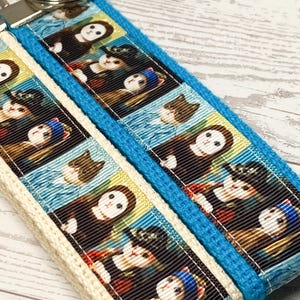 May include: A blue and cream-colored keychain wristlet featuring a repeating pattern of cat portraits. The wristlet has a silver-colored metal clasp and a blue woven border. The cat portraits are in a variety of poses and outfits.