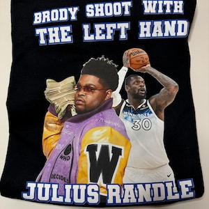 Pode incluir: Camiseta preta com uma estampa gráfica branca de dois jogadores de basquete. O texto "Brody Shoot With The Left Hand" é impresso em branco com um contorno azul. O texto "Julius Randle" é impresso em branco com um contorno azul. O gráfico apresenta um jogador de basquete vestindo uma jaqueta roxa e amarela e um jogador de basquete vestindo uma camisa branca com o número 30.