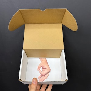 Pode incluir: Caixa de cartão aberta e vazia com interior branco. O interior da caixa apresenta uma imagem impressa de uma mão fazendo o gesto 'ok'. A caixa é segurada por uma mão contra um fundo preto.