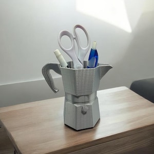May include: A gray, 3D-printed desk organizer shaped like a stovetop espresso maker. The organizer holds white scissors, a white marker, and a blue pen. The organizer sits on a wooden table.