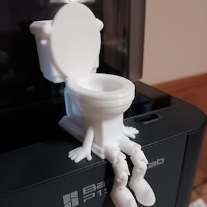 Puede incluir: Una figurita blanca de un inodoro con piernas y brazos, impresa en 3D. El inodoro está sentado sobre una superficie negra.