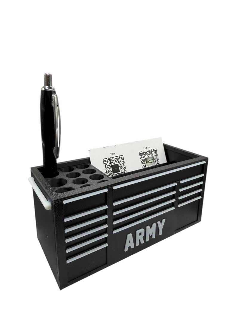 Op de afbeelding: Zwarte bureau-organizer in de vorm van een gereedschapskist, met witte accenten en het woord "ARMY" op de voorkant. Het heeft compartimenten voor pennen, visitekaartjes en een pen. Een zwarte en zilveren pen zit in de penhouder.