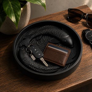 Puede incluir: Bandeja decorativa negra con dise&ntilde;o de peces koi. La bandeja contiene una cartera de cuero marr&oacute;n, llaves del coche y una llave. Gafas de sol y un reloj est&aacute;n sobre la superficie de madera. La bandeja tiene unos 30 cm de di&aacute;metro.