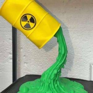 May include: A yellow barrel with a black and white radiation symbol is tilted, with green liquid pouring out. The liquid forms a puddle at the base. The sculpture is a unique decorative item.